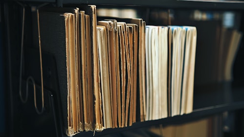 A collection of old, worn folders and files are neatly arranged on a dark shelf. The edges of the papers are yellowed, indicating age, and the surface of the shelf is a deep black, providing contrast to the light-colored documents.