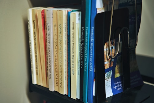 A collection of books organized on a shelf. The books are primarily reports on North Carolina pesticides, with publication years ranging from the early 1990s to the early 2000s. The books vary in color, including shades of yellow, orange, blue, and white. A black bookend supports them, and the titles are clearly visible.