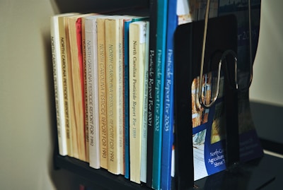 A collection of books organized on a shelf. The books are primarily reports on North Carolina pesticides, with publication years ranging from the early 1990s to the early 2000s. The books vary in color, including shades of yellow, orange, blue, and white. A black bookend supports them, and the titles are clearly visible.
