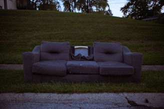 A worn-out grey couch with visible wear and sagging cushions is placed on a grassy curbside. Behind the couch, part of an old television set is visible, indicating possible disposal. The background includes a lush, green hill and trees with dim lighting suggesting early evening or late afternoon.