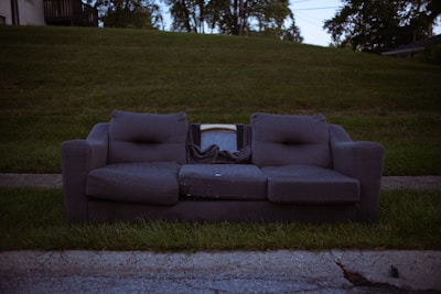 A worn-out grey couch with visible wear and sagging cushions is placed on a grassy curbside. Behind the couch, part of an old television set is visible, indicating possible disposal. The background includes a lush, green hill and trees with dim lighting suggesting early evening or late afternoon.