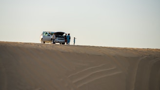 A group of people stands near an SUV with an open hood on a vast sand dune, suggesting a possible car breakdown or rest stop. The sand has smooth, wavy patterns, and the sky is clear.