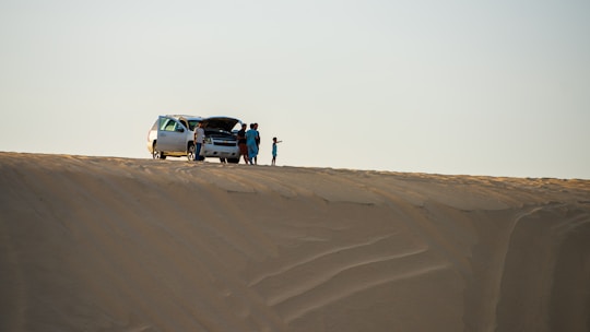 A group of people stands near an SUV with an open hood on a vast sand dune, suggesting a possible car breakdown or rest stop. The sand has smooth, wavy patterns, and the sky is clear.