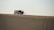 A group of people stands near an SUV with an open hood on a vast sand dune, suggesting a possible car breakdown or rest stop. The sand has smooth, wavy patterns, and the sky is clear.