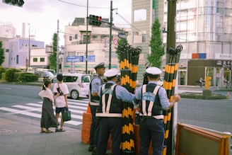 Several police officers stand near a crosswalk holding striped gates, possibly for traffic control. Pedestrians wait at the crosswalk, while cars are stopped on the other side. Urban buildings and traffic lights are visible in the background.