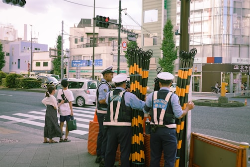 Several police officers stand near a crosswalk holding striped gates, possibly for traffic control. Pedestrians wait at the crosswalk, while cars are stopped on the other side. Urban buildings and traffic lights are visible in the background.