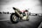 A sport motorcycle with a Honda logo is parked in an expansive, empty parking lot. The bike features a sporty design with a mix of colors including red, blue, and white. In the background, there are trees, light poles, and a modern overpass structure under a cloudy sky. The setting appears to be urban, with an industrial or commercial backdrop.