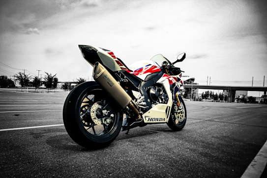 A sport motorcycle with a Honda logo is parked in an expansive, empty parking lot. The bike features a sporty design with a mix of colors including red, blue, and white. In the background, there are trees, light poles, and a modern overpass structure under a cloudy sky. The setting appears to be urban, with an industrial or commercial backdrop.