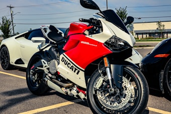 A red and white Ducati 899 Panigale motorcycle is parked next to a white sports car, with another dark-colored vehicle partially visible. The setting appears to be an outdoor parking lot with trees and a building in the background.
