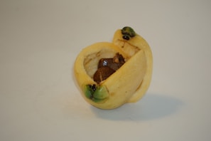 A close-up of a small pastry or dessert filled with a dark, possibly chocolate filling. It is wrapped in a light-colored dough, with green toppings at two corners, likely pistachios.