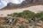 A scenic landscape featuring a rocky terrain with a small waterfall and a stream flowing through the foreground. Lush green bushes surround the water source, and two camping tents are set up on a sandy area. In the background, a majestic mountain range rises under a partly cloudy sky.