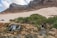 A scenic landscape featuring a rocky terrain with a small waterfall and a stream flowing through the foreground. Lush green bushes surround the water source, and two camping tents are set up on a sandy area. In the background, a majestic mountain range rises under a partly cloudy sky.