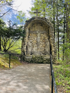 A stone grotto holding a crucifixion scene with three statues, including a central figure on a cross and two figures on either side. Surrounded by lush greenery, trees, and a stone pathway leading to the site. The top of the structure is crowned with a stone cross.