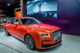 A luxury orange car with a sleek design is displayed indoors on a showroom floor. It features distinctive headlights, a prominent grille, and detailed wheels. Behind the car, there's a large screen displaying an artistic image of a car hood ornament.