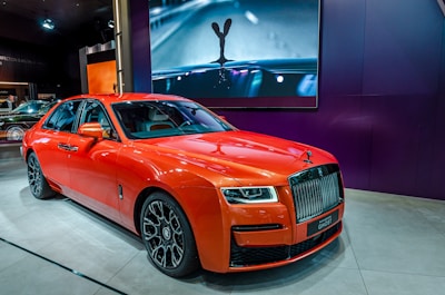 A luxury orange car with a sleek design is displayed indoors on a showroom floor. It features distinctive headlights, a prominent grille, and detailed wheels. Behind the car, there's a large screen displaying an artistic image of a car hood ornament.