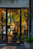 A cozy flower shop interior visible through a large glass door with gridded panes. The shop is filled with lush green plants and vibrant flowers in various colors, including white, purple, and yellow. A person is seen working at a table inside, surrounded by floral arrangements. Warm lighting creates a welcoming and serene atmosphere.