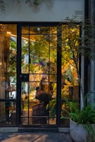 A cozy flower shop interior visible through a large glass door with gridded panes. The shop is filled with lush green plants and vibrant flowers in various colors, including white, purple, and yellow. A person is seen working at a table inside, surrounded by floral arrangements. Warm lighting creates a welcoming and serene atmosphere.