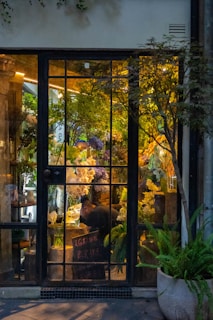 A cozy flower shop interior visible through a large glass door with gridded panes. The shop is filled with lush green plants and vibrant flowers in various colors, including white, purple, and yellow. A person is seen working at a table inside, surrounded by floral arrangements. Warm lighting creates a welcoming and serene atmosphere.