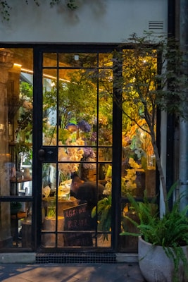 A cozy flower shop interior visible through a large glass door with gridded panes. The shop is filled with lush green plants and vibrant flowers in various colors, including white, purple, and yellow. A person is seen working at a table inside, surrounded by floral arrangements. Warm lighting creates a welcoming and serene atmosphere.