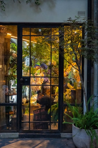 A cozy flower shop interior visible through a large glass door with gridded panes. The shop is filled with lush green plants and vibrant flowers in various colors, including white, purple, and yellow. A person is seen working at a table inside, surrounded by floral arrangements. Warm lighting creates a welcoming and serene atmosphere.