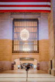 A dancer performs a high leg extension in a grand hall adorned with an American flag above and a large, ornate chandelier hanging in the center. The space features tall windows and intricate architectural design.