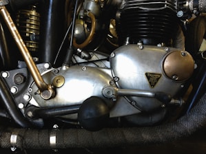 Close-up of a motorcycle engine with metal and brass components, showcasing intricate mechanical details and labels. The engine appears vintage, with visible bolts, levers, and textured surfaces.