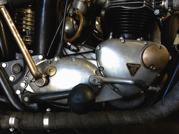 Close-up of a motorcycle engine with metal and brass components, showcasing intricate mechanical details and labels. The engine appears vintage, with visible bolts, levers, and textured surfaces.