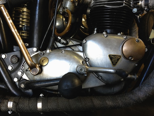 Close-up of a motorcycle engine with metal and brass components, showcasing intricate mechanical details and labels. The engine appears vintage, with visible bolts, levers, and textured surfaces.