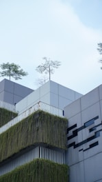 A modern building facade features sleek geometric designs and is adorned with green vertical gardens. Trees are planted atop the structure, adding a touch of nature. The walls are primarily composed of clean gray panels, and there are some artistic black patterns adding contrast.