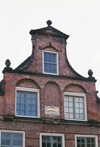 A historic brick building facade featuring symmetric window arrangements and intricate architectural detailing. The structure includes arched windows and a decorative plaque with a gold emblem and inscription, adding a vintage charm.