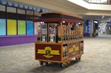 A candy cart filled with various jars of colorful candies, each with a dispenser. It is set in a shopping mall corridor with tiled floors and a partially visible storefront in the background.