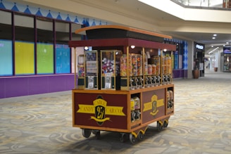 A candy cart filled with various jars of colorful candies, each with a dispenser. It is set in a shopping mall corridor with tiled floors and a partially visible storefront in the background.