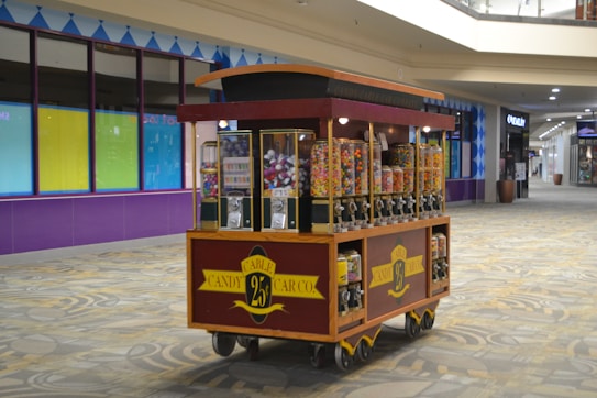 A candy cart filled with various jars of colorful candies, each with a dispenser. It is set in a shopping mall corridor with tiled floors and a partially visible storefront in the background.