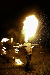 Two individuals are performing a fire-breathing act at night. The main performer is exhaling a large flame into the air, while another person in the background is holding and manipulating fire. The scene is illuminated by the bright flames, highlighting the performers against the dark surroundings.