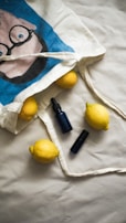 A cloth tote bag with a cartoon face print lies on a beige surface. Four lemons spill out of the bag along with two dark blue glass bottles, possibly for skincare or essential oils.