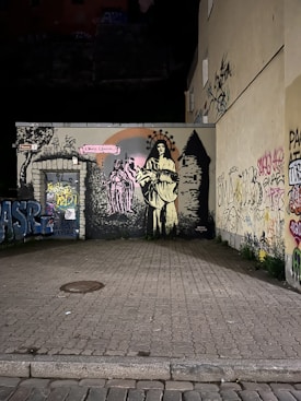 A detailed mural on a wall in an urban setting, featuring a prominent figure dressed in historical attire, holding an object with a crown-like design above their head. The mural is surrounded by graffiti and painted adjacent to another image of figures and a stone-like structure. The surrounding area has a paved ground and graffiti on nearby walls.