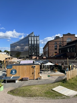 A modern glass building with a sleek design stands tall against a bright blue sky with scattered clouds. In the foreground, there is a small outdoor area featuring a wooden structure with various tables and chairs under umbrellas. The scene is lively and vibrant, with a mix of rustic and contemporary elements.