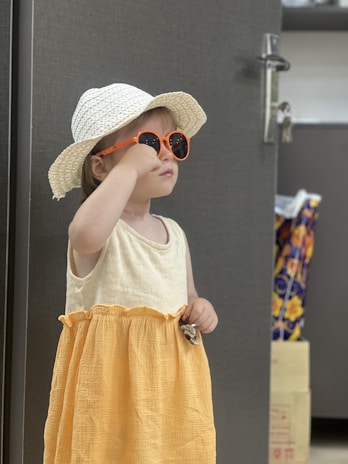 A young child wearing a light-colored sun hat and bright orange sunglasses stands indoors. The child is dressed in a sleeveless, two-toned dress with a creamy top and a mustard-yellow skirt. In the background are some cardboard boxes and a colorful bag.