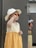 A young child wearing a light-colored sun hat and bright orange sunglasses stands indoors. The child is dressed in a sleeveless, two-toned dress with a creamy top and a mustard-yellow skirt. In the background are some cardboard boxes and a colorful bag.