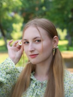 A young woman with long blonde hair is holding a small daisy near her face. She is wearing a floral-patterned blouse with light green and yellow accents. The background is a blurred green park setting, suggesting a natural and serene environment.