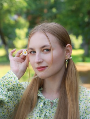 A young woman with long blonde hair is holding a small daisy near her face. She is wearing a floral-patterned blouse with light green and yellow accents. The background is a blurred green park setting, suggesting a natural and serene environment.