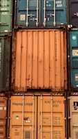 A stack of intermodal shipping containers featuring prominent brand labeling and identification numbers. The containers display various colors, primarily orange, blue, and green, and they are arranged in a grid-like formation. The surfaces show signs of weathering and rust, indicating frequent use and exposure to the elements.