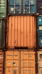 A stack of intermodal shipping containers featuring prominent brand labeling and identification numbers. The containers display various colors, primarily orange, blue, and green, and they are arranged in a grid-like formation. The surfaces show signs of weathering and rust, indicating frequent use and exposure to the elements.
