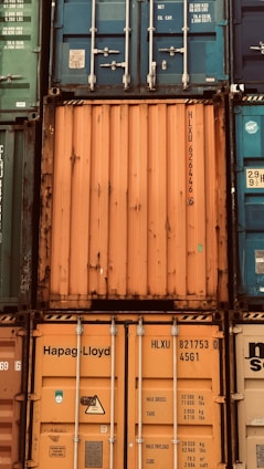 A stack of intermodal shipping containers featuring prominent brand labeling and identification numbers. The containers display various colors, primarily orange, blue, and green, and they are arranged in a grid-like formation. The surfaces show signs of weathering and rust, indicating frequent use and exposure to the elements.