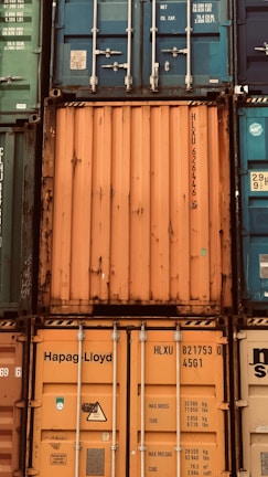 A stack of intermodal shipping containers featuring prominent brand labeling and identification numbers. The containers display various colors, primarily orange, blue, and green, and they are arranged in a grid-like formation. The surfaces show signs of weathering and rust, indicating frequent use and exposure to the elements.