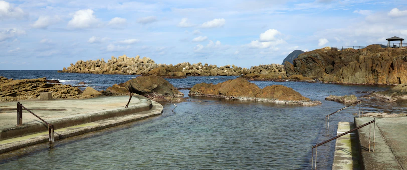 A coastal scene with a rocky shore and man-made barriers extending into the ocean. The foreground features a narrow channel filled with clear water, flanked by concrete paths with railings. In the distance, the horizon meets the sea under a sky scattered with clouds.