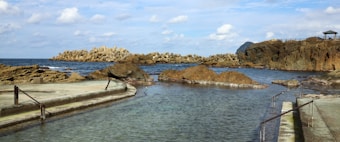 A coastal scene with a rocky shore and man-made barriers extending into the ocean. The foreground features a narrow channel filled with clear water, flanked by concrete paths with railings. In the distance, the horizon meets the sea under a sky scattered with clouds.