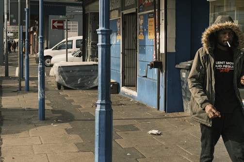 A man wearing a fur-lined hooded jacket is walking along a sidewalk in an urban setting, smoking a cigarette. The street is lined with blue painted poles and a mix of retail buildings. A white van is parked on the street alongside some wrapped furniture.