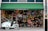 A storefront with a green awning labeled 'Hedonism Wines' is decorated with an array of colorful butterflies adorning the large glass windows. In front of the store, a white tricycle with a cart filled with more colorful decorations is parked. Inside the shop, warm lights create a cozy ambiance that is visible through the glass.