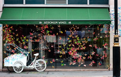 A storefront with a green awning labeled 'Hedonism Wines' is decorated with an array of colorful butterflies adorning the large glass windows. In front of the store, a white tricycle with a cart filled with more colorful decorations is parked. Inside the shop, warm lights create a cozy ambiance that is visible through the glass.